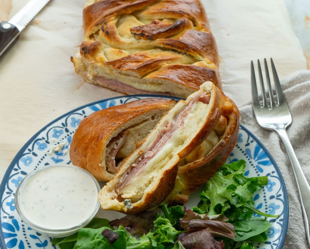 sliced baked ham and cheese sandwich on a plate with a side of ranch dressing fork off the the right side uncut portion of sandwich is on a cutting board behind plate