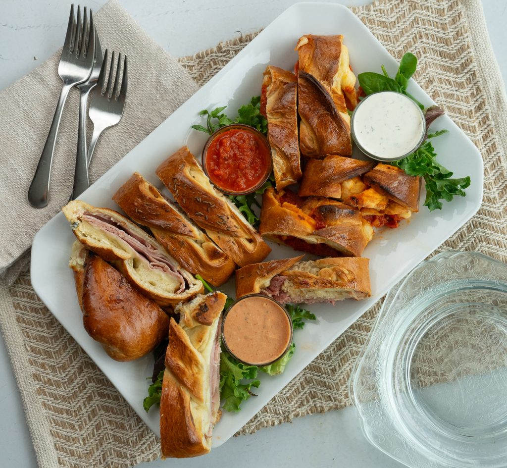 white platter with slices of baked sandwiches on it with small bowls of dips forks are placed with napkins in upper left hand corner and glass plates are stacked in lower right hand corner