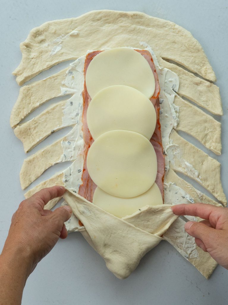 showing the bread braiding process of a baked ham and cheese sandwich. hands are folding up the bottom part of the sandwich