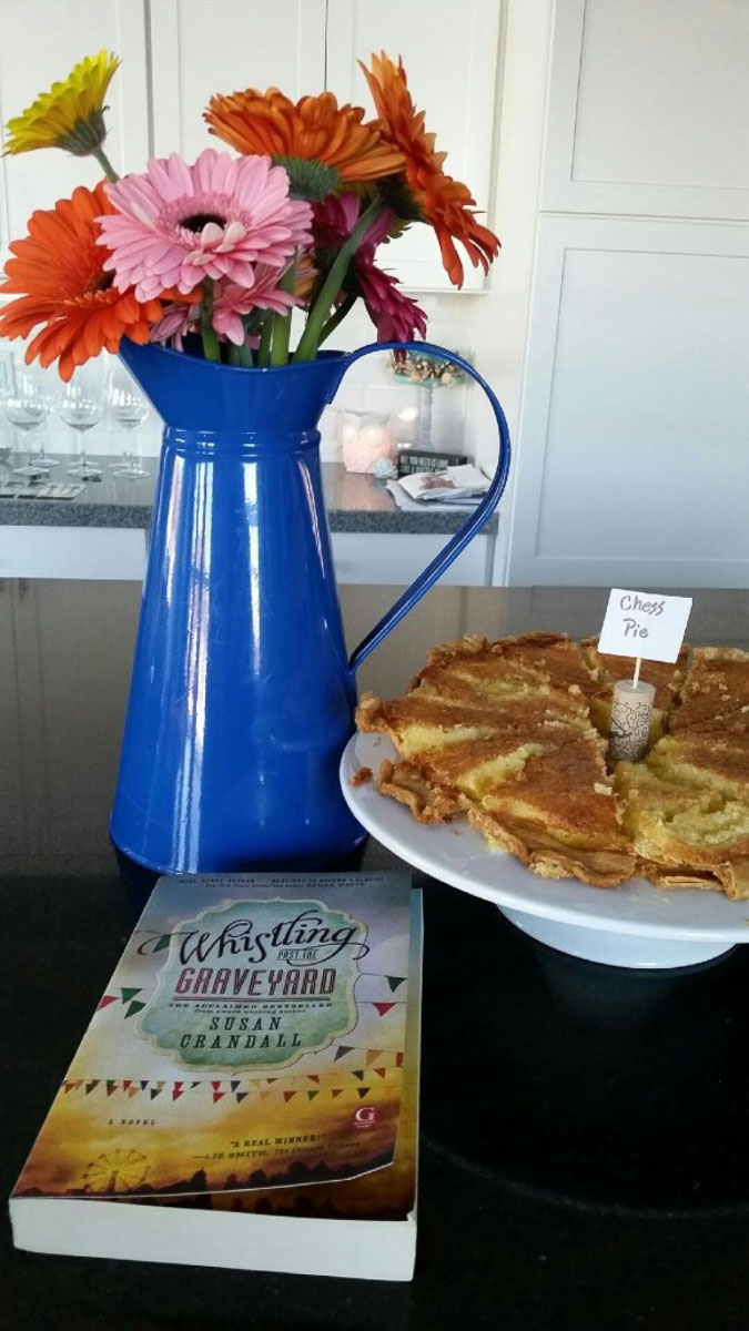 flours in a blue vase on a black counter top with a book and a chess pie also in the picture