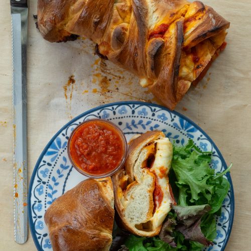 Baked Pepperoni Pizza sandwich on a cutting board with a plate with two slices of sandwich, dish of pizza sauce, and a green salad