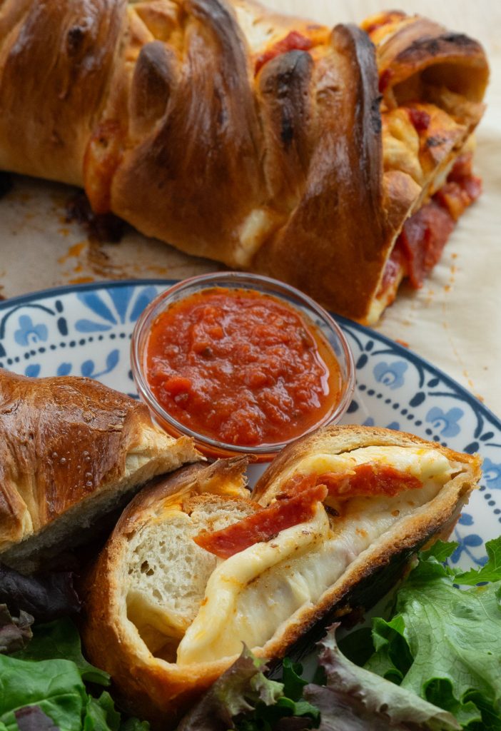 slice of baked pepperoni pizza sandwich on a plate with a dish of pizza sauce and an uncut sandwich on a cutting board above the plate 