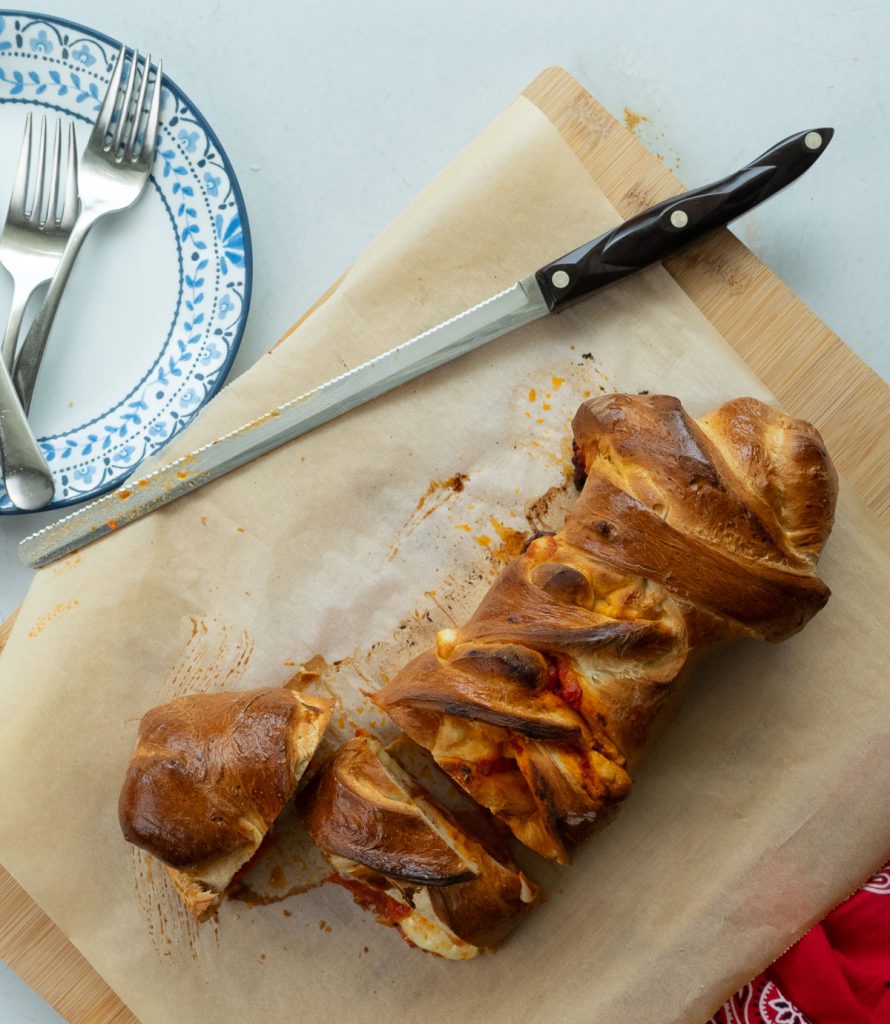 baked pizza sandwich on a cutting board with a bread knife plates and forks are set in the upper left hand corner