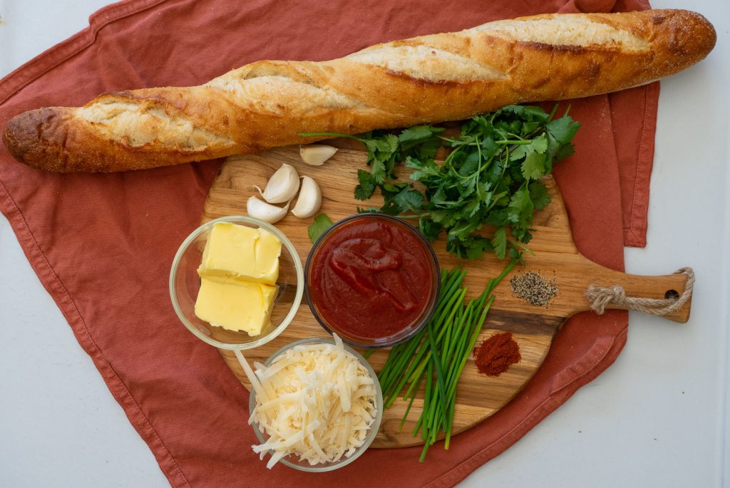 photo of ingredients needed for cilantro chili garlic bread on a round wood cutting board with a rust colored towel in background