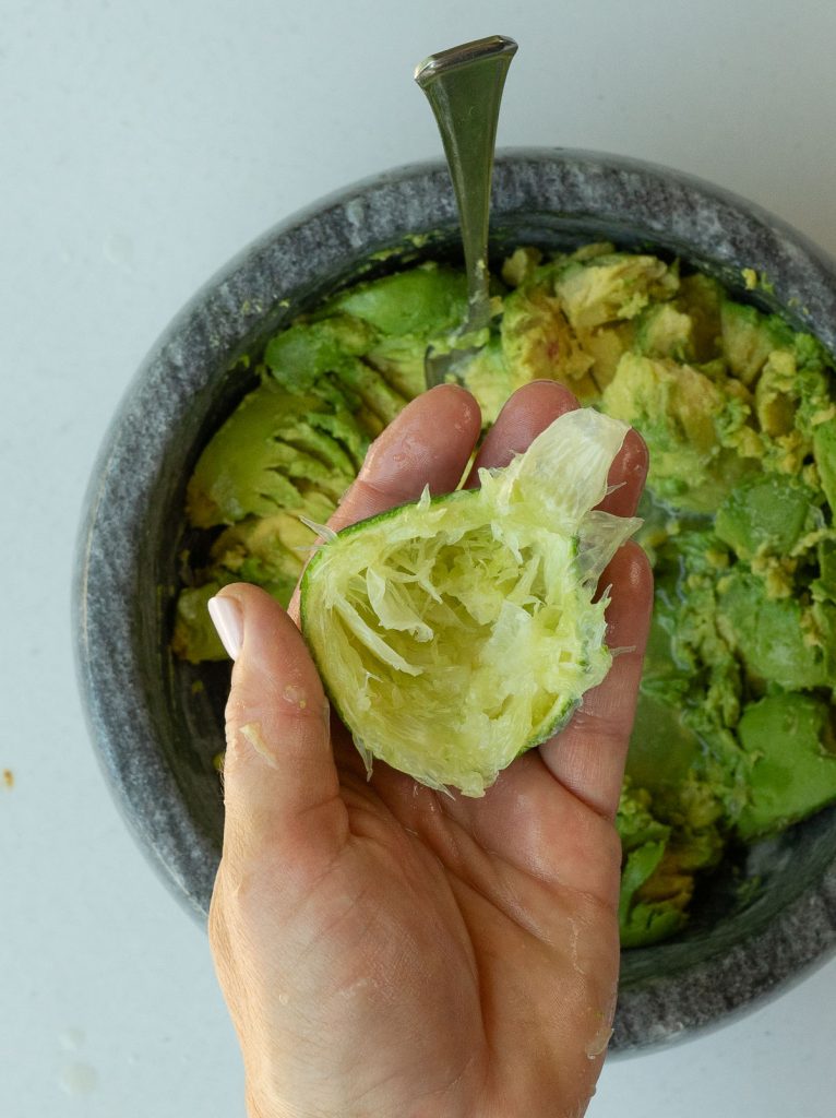 hand holding a fully squeezed lime over a bowl of smashed avocado