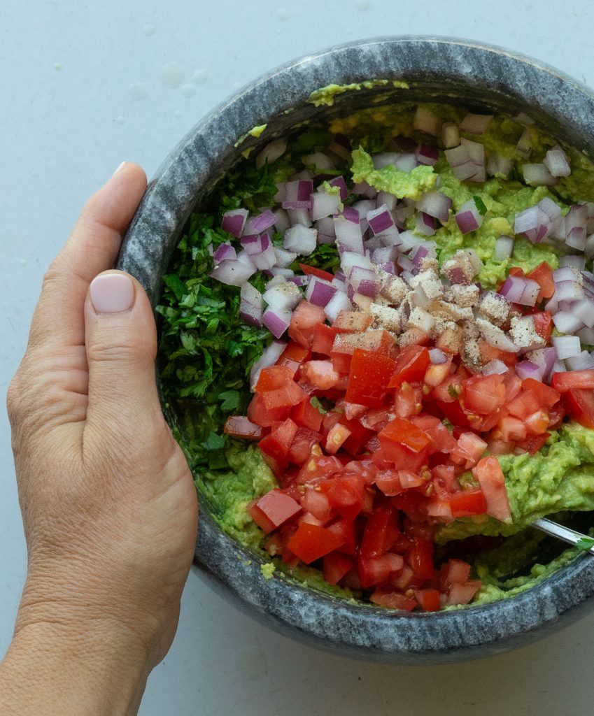 smashed avocados and lime juice in dark gray stone bowl with diced onion, cilantro and diced tomato and seasoning on top, hand on left side of bowl right hand is starting to stir