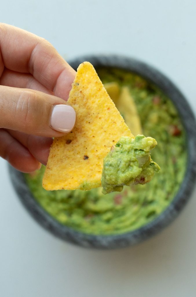 Corn tortilla chip with guacamole on it held by a hand with a bowl of guacamole in the background