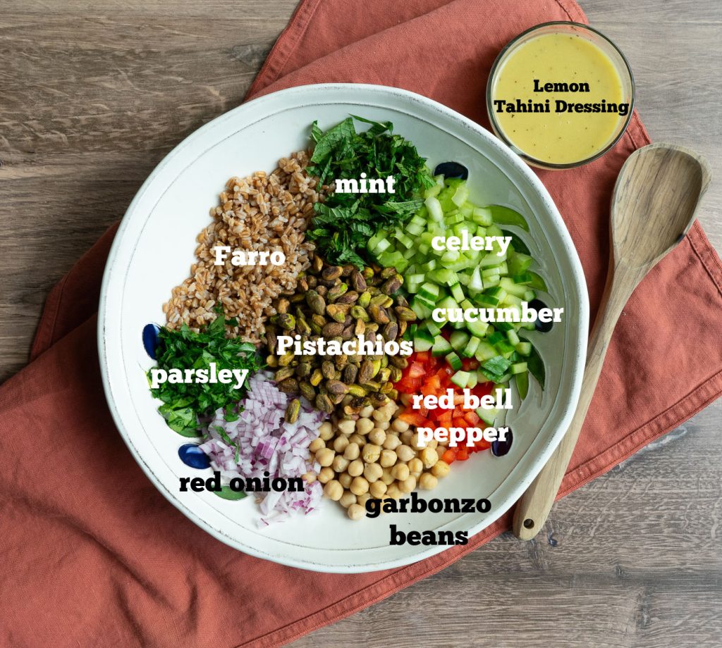 farro salad ingredients in a white  bowl with glass bowl of dressing at the top right corner with a wooden spoon laying beside the bowl