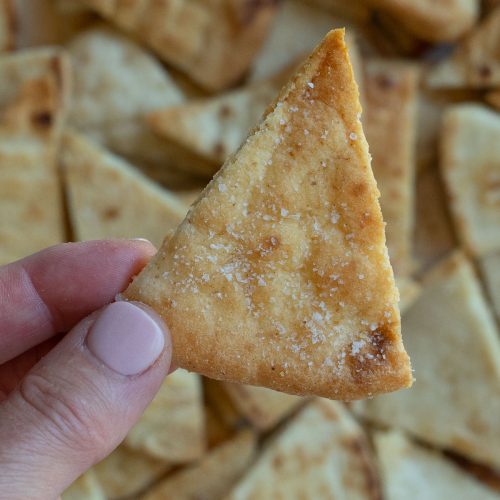 hand holding a single pita chip with a pan full of pita chips in the background