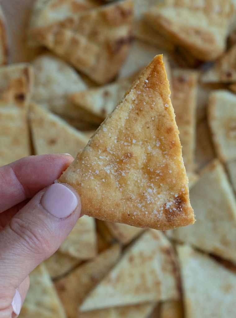 hand holding a single pita chip with a pan full of pita chips in the background