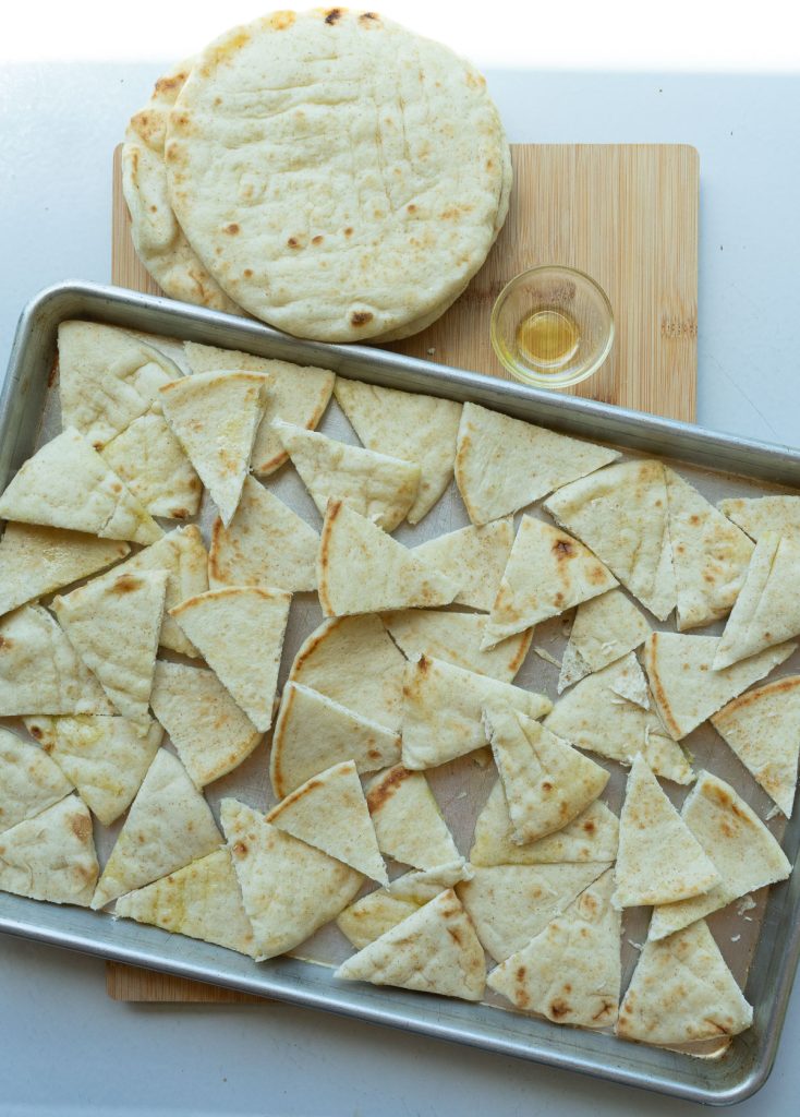 pita bread spread out in a single layer over a baking sheet that is sitting on a wood cutting board with whole pita bread in the upper left corner of photo and a small empty glass dish sitting next to pita bread 