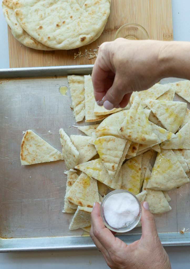 sprinkling salt over the pita bread that has been drizzled with olive oil on a baking sheet that is sitting on a wood cutting board with a stack of un cut pita bread in the upper left hand corner of the photo