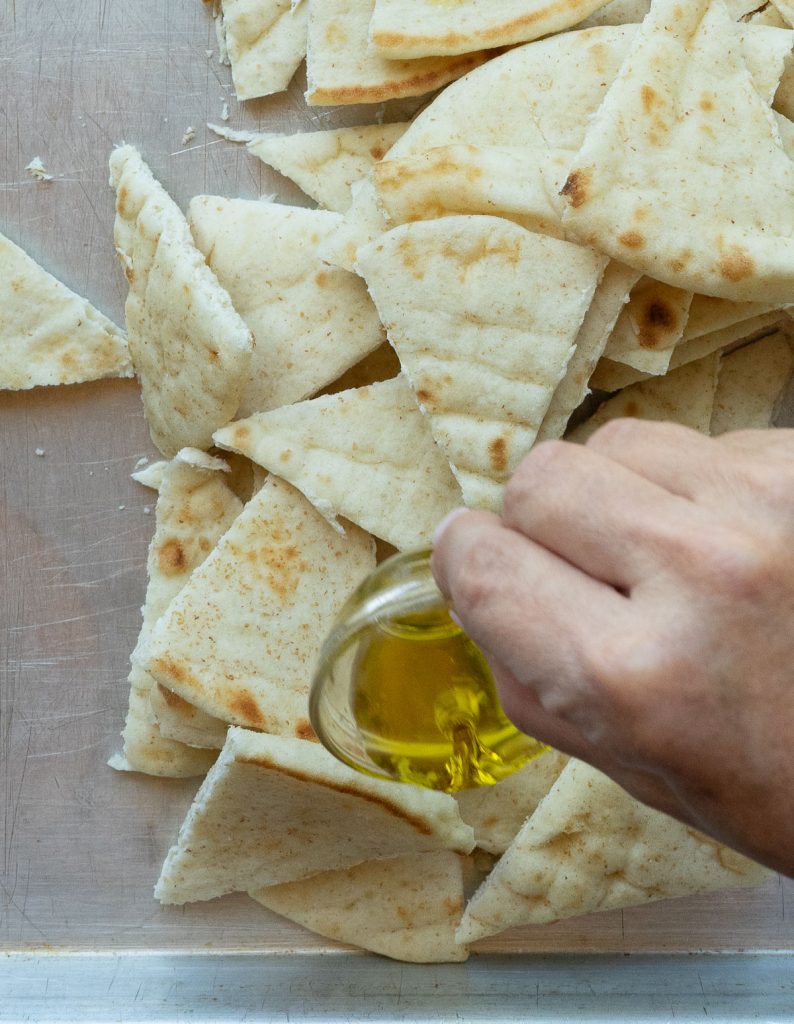 pile of pita bread triangles on a baking sheet with a hand pouring olive oil over the bread