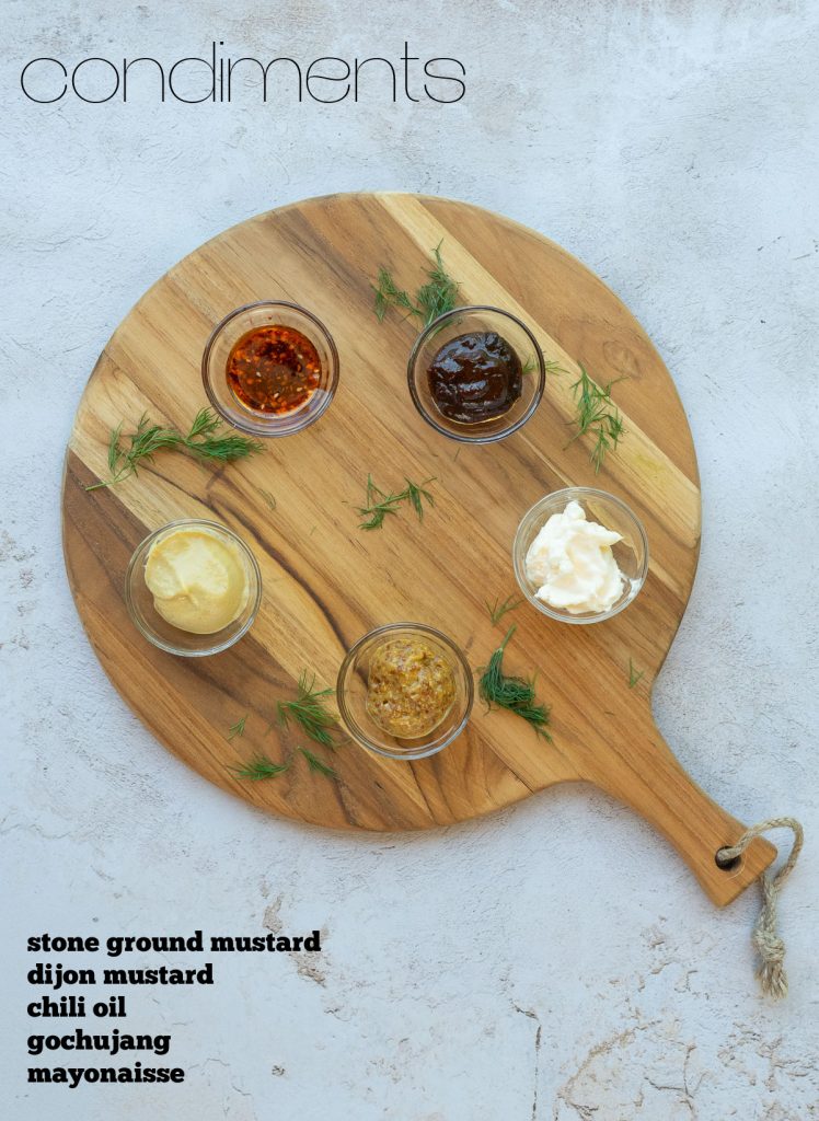 small glass dishes with various condiments placed on a round cutting board on a gray counter top with herbs sprinkled around