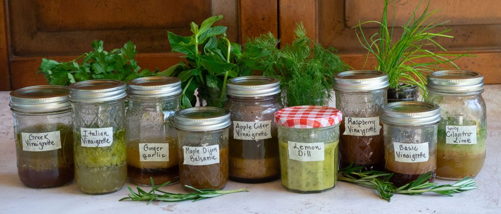 clear glass jars of varying heights are filled with different types of homemade vinaigrette lined up on table with labels on the jars fresh herbs are placed behind the jars. 