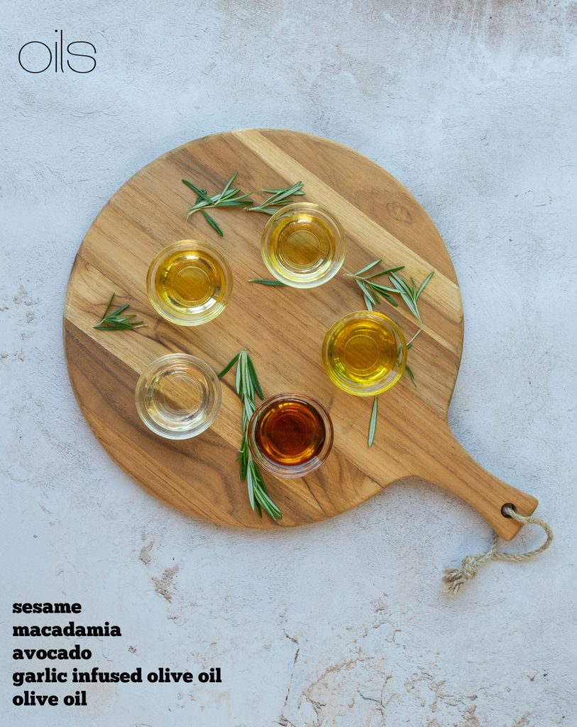 five different oils displayed in small glass dishes on a round wood cutting board on a gray counter top pieces of rosemary sprinkled around