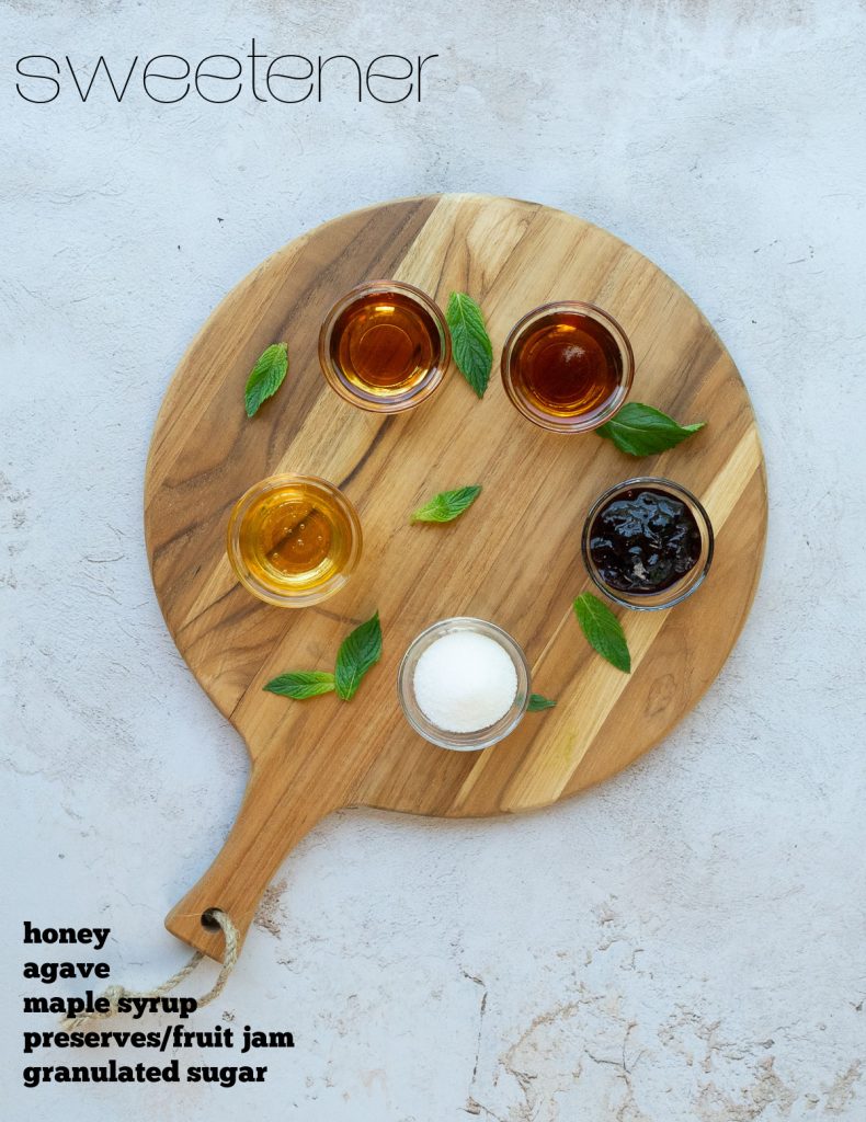 small glass dishes of sweeteners placed on a round wood cutting board on a gray counter top