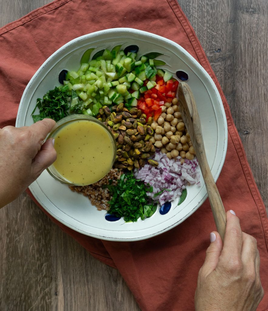 pouring lemon tahini dressing over farro salad ingredients in a large white bowl on wood counter top with a rust colored dish towel underneath the bowl