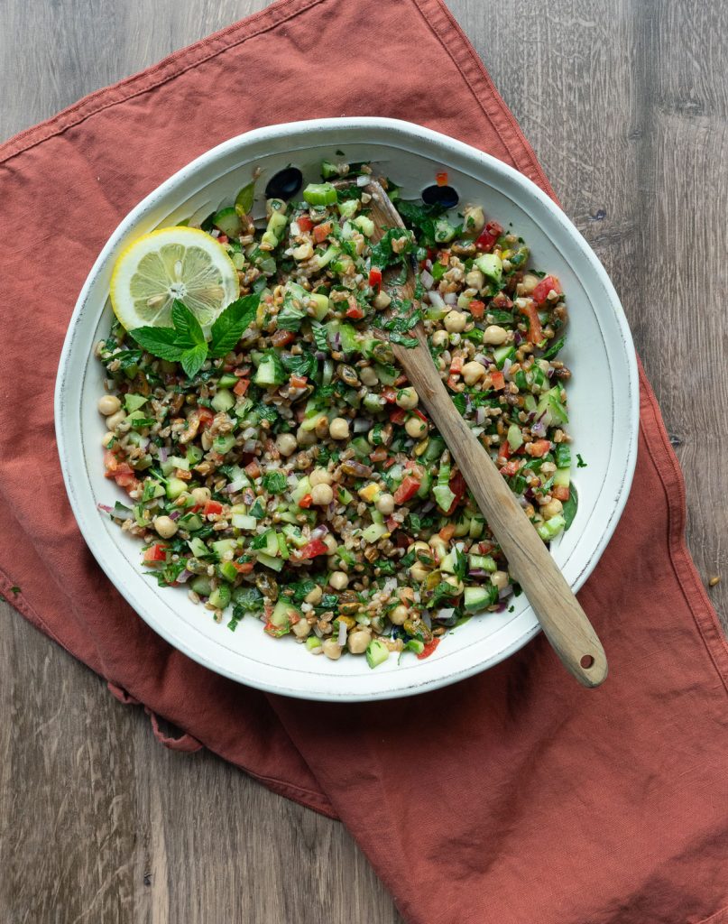 mixing up farro salad with a wooden spoon in a white bowl on wooden counter top with a rust colored napkin under the bowl 