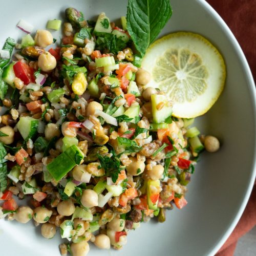 whole grain farro salad with lemon tahini dressing with lemon and mint garnish in a white bowl with a rust colored towel in background