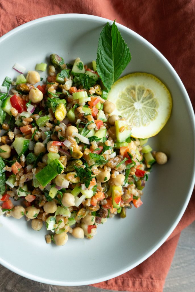 whole grain farro salad with lemon tahini dressing with lemon and mint garnish in a white bowl with a rust colored towel in background