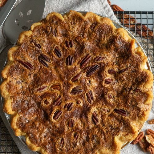 whole pecan pie settin on a cooling rack with pecans strewn on table pie server and knife off to the upper left side of the pie