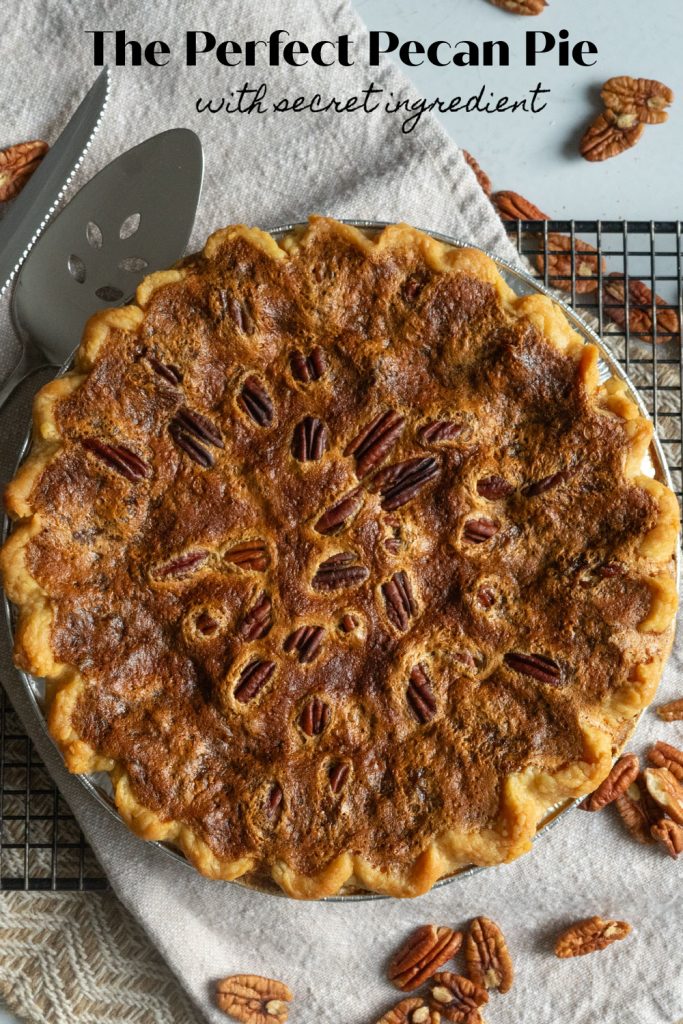 whole pecan pie settin on a cooling rack with pecans strewn on table pie server and knife off to the upper left side of the pie